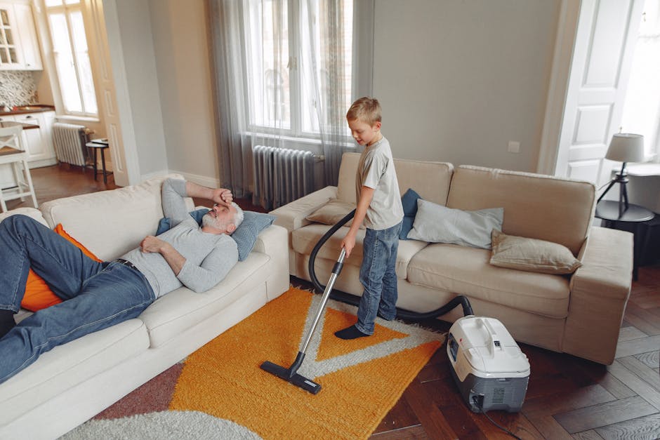 A young boy is vacuuming an orange and beige patterned carpet in a bright, well-lit living room, while an older man with gray hair and a beard lies on a white sofa, appearing relaxed. The room features light grey walls, large windows with sheer curtains, and decorative pillows on the sofa. A vacuum cleaner with a modern design is positioned on the hardwood floor near the boy, with its hose extended toward the carpet. The space appears clean, organized, and inviting, demonstrating domestic cleaning and surface maintenance in a residential setting, with emphasis on efficient deep cleaning using the vacuum cleaner from Carpet Cleaners SW4, as part of their surface cleaning and sanitisation services for Victorian homes in Clapham Common.