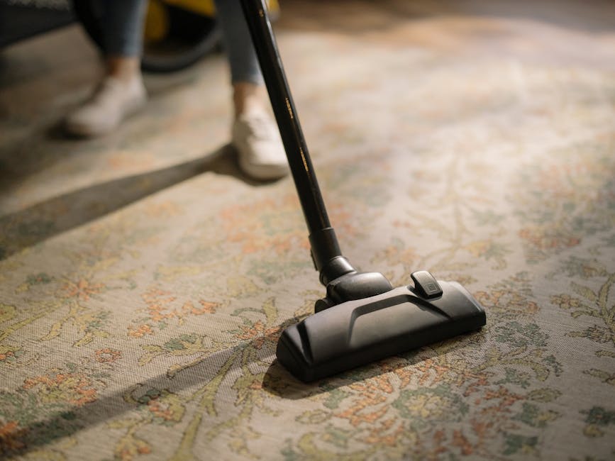 Close-up of a person's feet wearing beige shoes, operating a black vacuum cleaner with a slim handle, on a detailed patterned area rug with floral motifs in shades of green, beige, and orange. The scene takes place in a well-lit room, highlighting the surface cleaning process, with the vacuum's head positioned centrally on the rug. The background is slightly blurred, emphasizing the focus on the cleaning activity, which is part of a deep cleaning or maintenance task in a residential setting, consistent with the services offered by Carpet Cleaners SW4 for Victorian homes in Clapham Common.