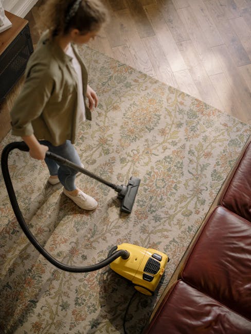 A young boy is vacuuming an orange and beige patterned carpet in a bright, well-lit living room, while an older man with gray hair and a beard lies on a white sofa, appearing relaxed. The room features light grey walls, large windows with sheer curtains, and decorative pillows on the sofa. A vacuum cleaner with a modern design is positioned on the hardwood floor near the boy, with its hose extended toward the carpet. The space appears clean, organized, and inviting, demonstrating domestic cleaning and surface maintenance in a residential setting, with emphasis on efficient deep cleaning using the vacuum cleaner from Carpet Cleaners SW4, as part of their surface cleaning and sanitisation services for Victorian homes in Clapham Common.