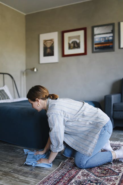 A woman with brown hair tied back, wearing a light blue striped shirt and jeans, is kneeling on a patterned rug while using a blue cloth to clean the dark wooden floor in a living room. The room featu