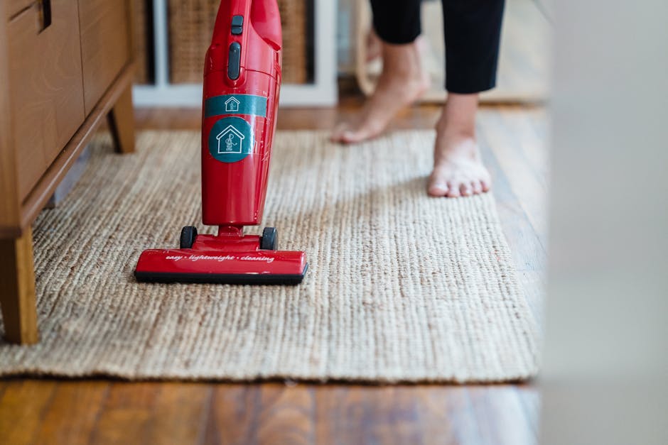 A woman with brown hair tied back, wearing a light blue striped shirt and jeans, is kneeling on a patterned rug while using a blue cloth to clean the dark wooden floor in a living room. The room featu