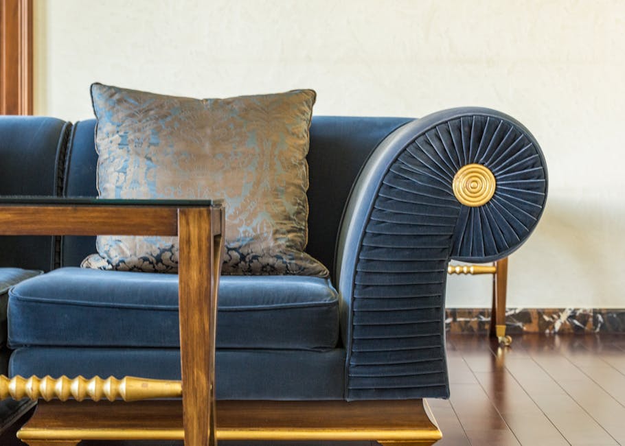 A white upholstered sofa with wooden legs situated in a bright, minimalist room with white walls and flooring. A beige throw blanket is casually draped over one armrest and the seat, adding a soft tex